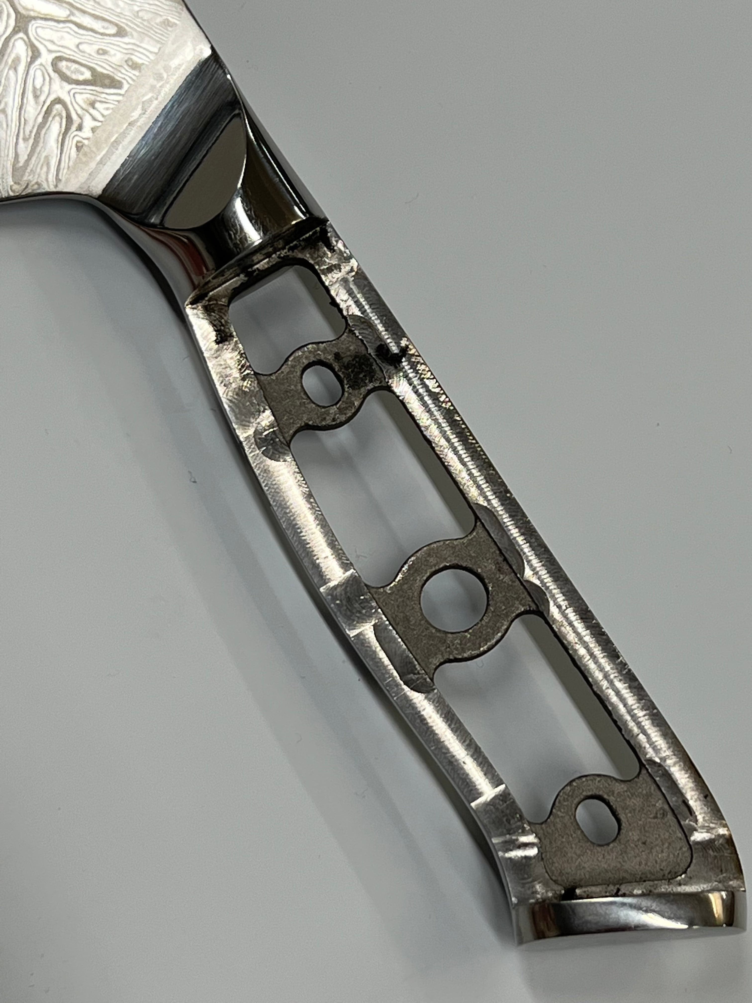 Nærbilde av en polert Sild Santoku - VG10 Damask Kokkekniv 7" håndtak laget av metall. Håndtaket har en åpen rammedesign med en rekke store og små geometriske utskjæringer, som avslører dens intrikate struktur. Metallet har en reflekterende overflate, og bakgrunnen er en jevn, lys grå farge.