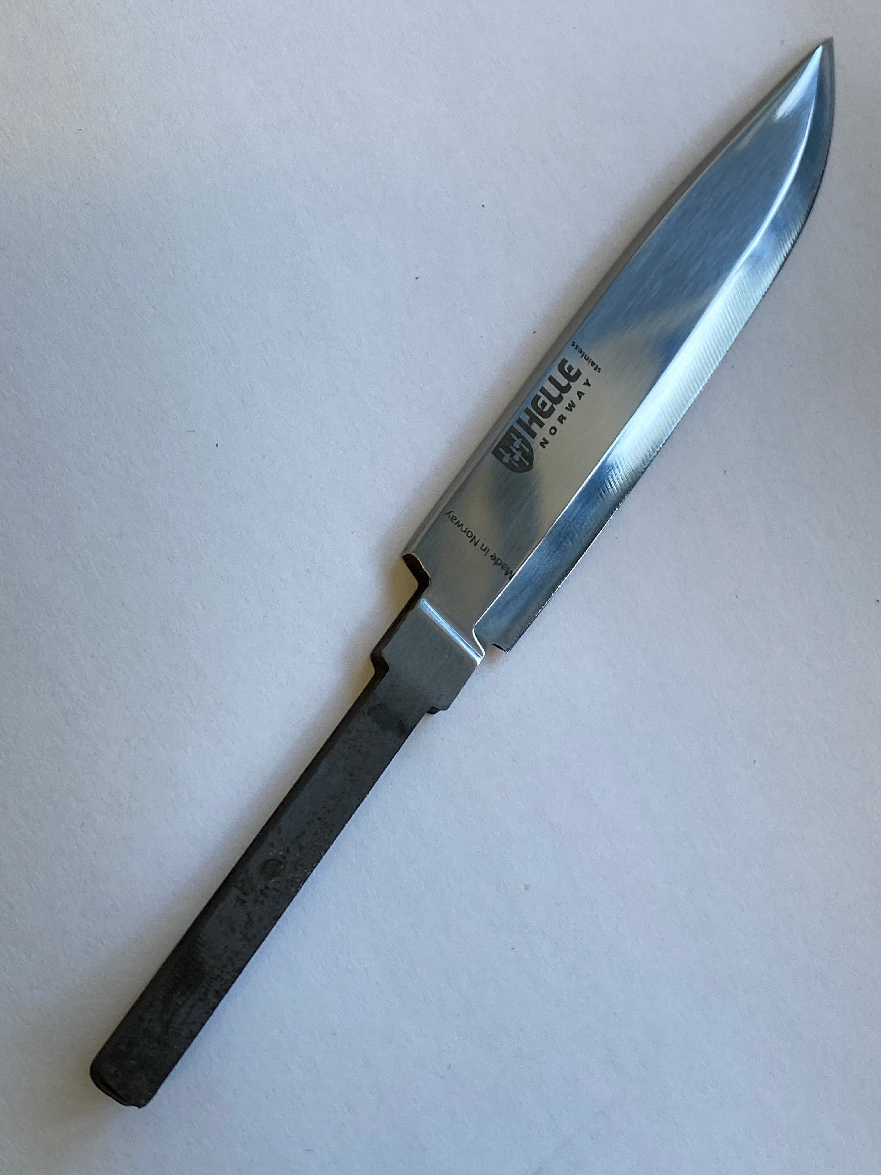 En delvis ferdigbehandlet kniv med et skinnende, polert Sandvik 12C27-blad og et uraffinert, teksturert håndtak. Bladet, merket med merkenavnet "Helle" og "Norge", fremkaller kvaliteten til Helle Speiderkniven 04. Bakgrunnen er en vanlig, lys overflate.