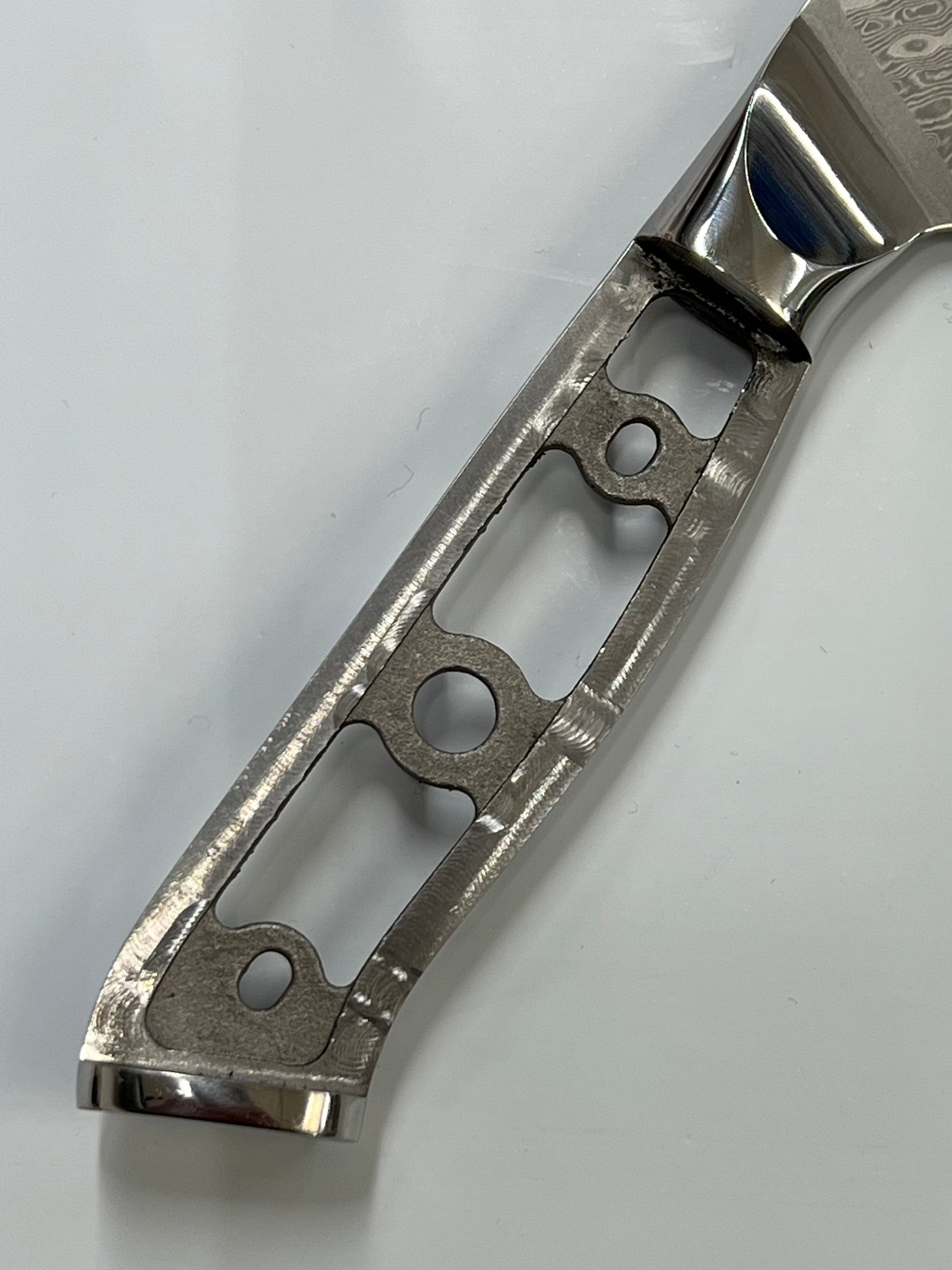 Nærbilde av et knivskaft designet med en intrikat åpen rammestruktur. Håndtaket har flere utskårne seksjoner, som danner et særegent geometrisk mønster, og er laget av polert rustfri damaskmetall. De glatte kantene og det minimalistiske utseendet understreker håndverket og den moderne designen til dette Herring Santoku - VG10 Damask Kokkekniv 7" håndtaket.