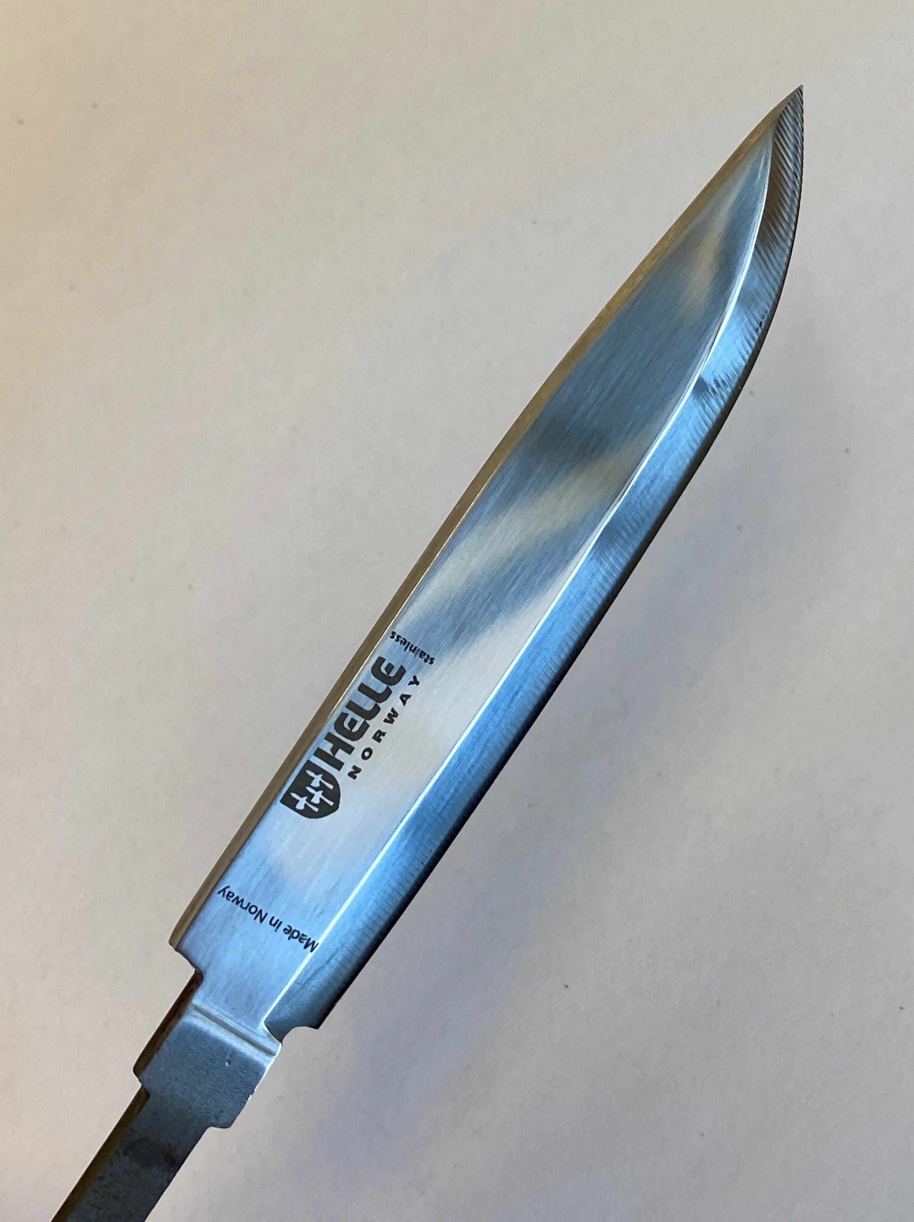 En närbild av ett metalliskt knivblad mot en slät bakgrund. Bladet, tillverkat av Sandvik 12C27-stål, är präglat med varumärket "Helle" och andra markeringar. Denna Helle Scout-kniv 04 har en vass, rak egg, spetsig spets, synlig sliplinje och en lätt reflekterande yta.