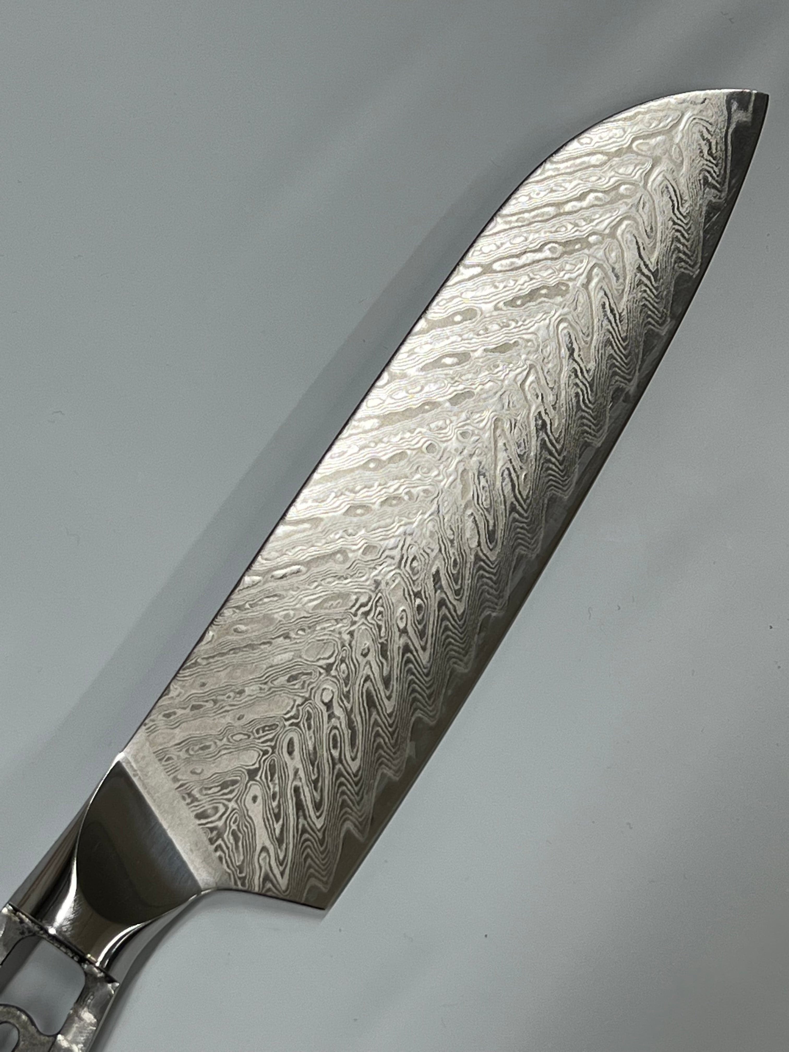 Nærbilde av en slank, høykvalitets Sild Santoku - VG10 Damask Kokkekniv 7" blad med et bølget, intrikat rustfrit damaskmønster. VG10 eggstålbladet er skarpt og reflekterende, og viser dets håndverk og design mot en nøytral bakgrunn. Håndtaket er delvis synlig, laget av polert metall.